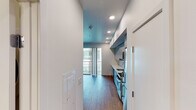 This single-floor unit showcases a contemporary design with warm hardwood flooring and a clean, minimalist aesthetic throughout. The open layout connects the kitchen and living areas seamlessly, creating an efficient and functional flow.

The kitchen is a standout feature, lined with high-gloss, powder-blue cabinetry extending from floor to ceiling along a single wall. White countertops contrast the cabinetry, complemented by stainless steel appliances including a gas range, built-in microwave, and dishwasher. Recessed lighting illuminates the space effectively.

The living area is bright and open, with crisp white walls and natural light pouring in through large floor-to-ceiling sliding glass doors that lead to a private balcony with metal railing. The warm wood flooring carries throughout, maintaining a cohesive visual continuity.

A connecting hallway provides access to additional rooms, with recessed lighting and white walls maintaining the clean design language. The bathroom features a white vanity with an undermount sink, a large wall mirror, white subway tile, a combined tub and shower, and matching hardwood flooring, all presented in a bright, functional arrangement.