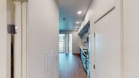 This single-floor unit showcases a contemporary design with warm hardwood flooring and a clean, minimalist aesthetic throughout. The open layout connects the kitchen and living areas seamlessly, creating an efficient and functional flow.

The kitchen is a standout feature, lined with high-gloss, powder-blue cabinetry extending from floor to ceiling along a single wall. White countertops contrast the cabinetry, complemented by stainless steel appliances including a gas range, built-in microwave, and dishwasher. Recessed lighting illuminates the space effectively.

The living area is bright and open, with crisp white walls and natural light pouring in through large floor-to-ceiling sliding glass doors that lead to a private balcony with metal railing. The warm wood flooring carries throughout, maintaining a cohesive visual continuity.

A connecting hallway provides access to additional rooms, with recessed lighting and white walls maintaining the clean design language. The bathroom features a white vanity with an undermount sink, a large wall mirror, white subway tile, a combined tub and shower, and matching hardwood flooring, all presented in a bright, functional arrangement.