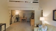 This two-story residence makes a striking first impression with its soaring double-height ceilings that flood the main living area with a sense of openness and volume. The ground floor features an open-plan layout where the living and dining areas flow seamlessly into one another, unified by warm neutral-toned carpet and soft, ambient lighting. Pendant lights hang gracefully above the dining area, which is centered around a wood dining table and chairs, complemented by a matching sideboard and decorative accents. Lush indoor plants add a natural, welcoming touch to the space. The living area is finished with comfortable seating and stylish furnishings that reinforce the warm, inviting atmosphere. A staircase leads to the upper level, where a loft-style space overlooks the main floor below through an open railing, creating a dynamic visual connection between the two levels. The upper floor benefits from natural light, with large windows visible from below. The overall design balances functionality with an airy, spacious ambiance, making excellent use of vertical space throughout. Neutral wall tones and consistent flooring choices tie the entire home together in a cohesive and comfortable aesthetic.