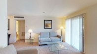 This furnished single-floor residence at Hunters Glen offers a well-organized layout designed for comfortable everyday living. The living room serves as the central gathering space, featuring plush wall-to-wall carpeting, neutral-toned walls, and a large sliding glass door dressed with vertical blinds that allows ample natural light to fill the room. The space is furnished with a light blue sofa, glass coffee table, and accent lamps, creating a clean and inviting atmosphere. A second seating area near the entryway provides additional living space, complemented by a built-in closet for convenient storage.

The kitchen is efficiently designed with warm wood cabinetry lining both walls, light countertops, a stainless steel sink, and tile flooring. It is equipped with a refrigerator, electric range, and dishwasher, offering full functionality.

The bedroom is a comfortably sized retreat with soft carpeting, a window overlooking greenery, and a dedicated closet with built-in shelving and hanging space. The bathroom features tile flooring, a wood-toned vanity with a light countertop, a full tub and shower combination, and a large mirror above the sink, maintaining a clean and practical design throughout.