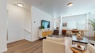 This well-appointed three-story residence showcases a cohesive mid-century modern aesthetic across all levels, with bright white walls, warm wood-tone flooring, and an abundance of natural light filtering through slatted blinds throughout.

The main living area is an open, inviting space anchored by a media console and wall-mounted TV, with room to accommodate both a comfortable seating area and a dedicated dining space. The dining area features a solid wood trestle table set beneath generous windows that bathe the room in natural light. Adjacent to the living and dining areas, the kitchen stands out with dark espresso cabinetry, stainless steel appliances, a subway tile backsplash, and a large center island with a white quartz countertop and seating — ideal for casual dining or entertaining. Pendant lighting adds a stylish focal point above the island.

A secondary living area on another level offers additional lounge space with warm tones and curated artwork. The bedrooms are well-proportioned and carpeted, each offering a calm, restful atmosphere with natural light and tasteful décor. The bathrooms are clean and functional, featuring full soaking tub and shower combinations with white surround walls, modern fixtures, and wood-look flooring. Stairs connect all three levels, making the layout practical and easy to navigate.