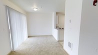 This single-floor unit presents a straightforward and functional layout designed for comfortable everyday living. The living area is finished with plush wall-to-wall carpeting in a neutral tone, creating a warm and understated aesthetic throughout. Bright white walls and a flush ceiling light fixture keep the space feeling clean, airy, and well-lit.

A defining feature of this unit is the open pass-through kitchen, which connects seamlessly to the living area. The kitchen includes white cabinetry, a countertop with a built-in sink, and a practical half-wall partition that maintains a sense of openness while defining the cooking space. This design allows for easy interaction between the kitchen and living areas.

Natural light filters into the living space through a large sliding glass door framed by floor-length white curtains, adding brightness and a sense of openness to the room. A wall-mounted heating unit is conveniently located near the base of the wall, ensuring year-round comfort.

The overall ambiance of the space is clean, minimal, and move-in ready, with a neutral color palette that provides a versatile backdrop for a variety of furnishing styles. The efficient layout maximizes usable space, making it a practical and welcoming environment.