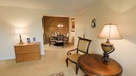 This single-floor residence offers a cohesive and comfortable living environment with a classic, traditional design aesthetic. The living area is furnished with elegant wood-accented pieces, including a round wooden table and cushioned armchair, creating an inviting atmosphere anchored by soft, neutral carpeting that flows throughout the space. A wooden dresser adds functional storage while complementing the warm tones of the interior. The space opens directly into a formal dining area, which serves as a standout feature of the home. A dramatic exposed brick accent wall anchors the dining room, lending a sense of character and timeless charm. The dining area is centered around a round table surrounded by classic dark-framed chairs with upholstered seats, and illuminated by an elegant chandelier that casts warm light over the room. Framed artwork adorns the brick wall, adding a cultivated, curated touch to the space. A nearby doorway with a closet suggests thoughtful storage solutions integrated into the layout. The consistent use of soft carpeting, warm lighting, and traditional furnishings throughout creates a harmonious and welcoming ambiance, making this residence an ideal blend of comfort and classic style.