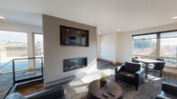 Located at Bridgeport Way W in University Place, Washington, this single-floor communal lounge space offers a contemporary design with a carefully curated layout. The open-plan interior is anchored by a striking feature wall housing a recessed linear fireplace beneath a wall-mounted flat-screen television, creating a sophisticated focal point for the main seating area. Dark leather armchairs and sofas are arranged around a round coffee table atop textured area rugs, providing a comfortable and stylish gathering space. Recessed lighting throughout the clean white ceilings supplements the generous natural light that filters in through large, dark-framed windows, offering views of the surrounding Pacific Northwest landscape. A dedicated kitchenette area along one wall includes dark cabinetry, a sink, and appliances, supporting event and gathering needs. Large-format tile flooring transitions to patterned carpet in the seating zones, adding visual interest. A secondary dining area with table and chairs sits adjacent to the windows, and glass-paneled entry doors connect to additional spaces. The overall ambiance is polished and modern, balancing function with a welcoming, upscale aesthetic.