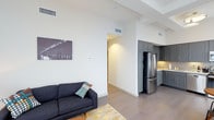 This single-floor space showcases a thoughtfully designed open-concept layout that seamlessly connects the living area and kitchen. The living space features soaring ceilings with exposed beams and ductwork, contributing to a spacious, airy atmosphere. Neutral white walls provide a clean backdrop, complemented by smooth, wide-plank flooring that runs throughout the unit.

The kitchen is outfitted with sleek gray cabinetry extending to the ceiling for generous storage, stainless steel appliances including a full-size refrigerator, and a warm tile backsplash that adds a subtle textural element to the space. The cabinetry's modern profile and muted tone create a sophisticated, contemporary look.

The living area flows naturally from the kitchen, offering an open and versatile space. A wide hallway leads further into the unit, suggesting additional rooms beyond the main living and kitchen zone. Recessed lighting and ceiling-mounted HVAC vents are integrated cleanly into the design, maintaining the minimalist aesthetic. Overall, the space balances functionality and modern style with a neutral color palette, high-quality finishes, and an efficient, well-organized floor plan.