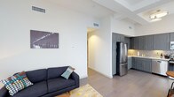 This single-floor space showcases a thoughtfully designed open-concept layout that seamlessly connects the living area and kitchen. The living space features soaring ceilings with exposed beams and ductwork, contributing to a spacious, airy atmosphere. Neutral white walls provide a clean backdrop, complemented by smooth, wide-plank flooring that runs throughout the unit.

The kitchen is outfitted with sleek gray cabinetry extending to the ceiling for generous storage, stainless steel appliances including a full-size refrigerator, and a warm tile backsplash that adds a subtle textural element to the space. The cabinetry's modern profile and muted tone create a sophisticated, contemporary look.

The living area flows naturally from the kitchen, offering an open and versatile space. A wide hallway leads further into the unit, suggesting additional rooms beyond the main living and kitchen zone. Recessed lighting and ceiling-mounted HVAC vents are integrated cleanly into the design, maintaining the minimalist aesthetic. Overall, the space balances functionality and modern style with a neutral color palette, high-quality finishes, and an efficient, well-organized floor plan.