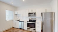 This efficiently designed single-floor unit offers a thoughtfully laid-out interior comprising a kitchen, living room, and full bathroom, all finished with a clean, modern aesthetic. Warm wood-look flooring flows consistently throughout the space, complementing the bright white walls and recessed lighting that give the interior a fresh, airy feel.

The kitchen is a standout feature, equipped with a full suite of stainless steel appliances including a refrigerator, electric range, over-the-range microwave, and dishwasher. Sleek white cabinetry extends from counter height to the ceiling, maximizing storage, while a white subway tile backsplash and light quartz-style countertops add a polished, contemporary touch. Large windows fitted with horizontal blinds allow natural light to filter in and offer views of surrounding greenery, with access to a small outdoor balcony area.

The open living area is bright and versatile, featuring recessed lighting and a Murphy-style cabinet that adds functional storage without occupying additional floor space. The textured ceiling and clean white walls maintain the unit's modern, minimalist character.

The bathroom is fully appointed with a single-sink vanity topped with a white surface, a large mirror with overhead lighting, a toilet, and a full soaking tub with shower and built-in shelving, all finished in crisp white tones with coordinating wood-look flooring.