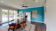 This single-floor communal space is thoughtfully designed to blend comfort and style in an open, welcoming layout. Bold teal accent walls serve as a defining visual element, contrasting with soft gray walls and warm dark hardwood flooring that runs throughout. Crown molding and recessed lighting add architectural refinement, while ceiling fans contribute both function and a modern aesthetic.

The lounge area centers around a stone-surround fireplace topped with a wall-mounted television, creating a cozy focal point for relaxation. A plush sofa with colorful accent pillows is arranged atop a teal-accented area rug, complemented by a sleek round ottoman. Glass French doors open directly to a poolside patio, flooding the space with natural light and providing seamless indoor-outdoor connectivity with views of the surrounding pool area.

Adjacent to the lounge, a round glass-top dining table with mid-century-style wood chairs offers a casual gathering spot. A bar-height counter with dark upholstered stools lines one wall, ideal for socializing or informal meetings. Decorative artwork, including abstract paintings and a textured wall panel, adds personality throughout. A workstation with computer equipment provides a functional nook within the space.