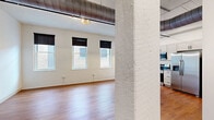 Located at 219 N Columbus St in Lancaster, Ohio, this 2-bedroom, 1-bathroom unit offers a thoughtfully updated interior with a blend of industrial character and modern finishes. The open living area features warm hardwood flooring, exposed white-painted brick walls, and a beadboard ceiling with visible ductwork and piping that lend a distinct loft-style aesthetic. Multiple windows along the main living space flood the interior with natural light and provide views of the surrounding streetscape.

The kitchen is a standout feature, equipped with stainless steel appliances including a refrigerator, electric range with over-the-range microwave, and a deep undermount sink with a matte black faucet. White shaker-style cabinetry with black hardware lines the walls and pairs with a central island offering additional counter space and pendant lighting overhead.

The two bedrooms are carpeted for comfort and each benefit from natural light through individual windows with roller shades. The primary bedroom offers a generous footprint with a slatted ceiling detail adding visual interest.

The bathroom showcases a clean, contemporary design with hexagonal tile flooring, a sage-toned vanity with white countertop, a framed mirror, and black accent hardware throughout. This unit presents a cohesive mix of modern updates within a building rich with historic character.