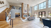 Located at 527 Old Bridge Turnpike in East Brunswick, NJ, this two-story clubhouse is a thoughtfully designed communal space that blends contemporary architecture with upscale, functional amenities. The main floor is anchored by a dramatic double-height great room with soaring ceilings, wide-plank hardwood flooring, and floor-to-ceiling windows that flood the space with natural light and offer views of the surrounding outdoors. A striking marble-clad fireplace serves as the room's centerpiece, flanked by warm wood columns and complemented by designer branch-style chandeliers. Multiple seating arrangements invite both casual lounging and social gatherings. Adjacent to the main living area is a sleek kitchen and bar space featuring an extended concrete countertop, dark cabinetry, pendant lighting with gold-toned fixtures, a built-in coffee station, and a media wall — ideal for entertaining. A dedicated dining room offers banquette seating with textured accent walls and ambient pendant lighting, creating an intimate setting. A second living area on the main floor includes a built-in shelving unit with a TV, additional lounge seating, and ample natural light. The upper level overlooks the great room via a modern cable-railing mezzanine and houses a professional conference room with a marble-top table, blue upholstered chairs, and refined finishes throughout.