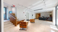 Located at 7 New York Ave NE in Washington, DC, this contemporary building lobby offers a refined and welcoming first impression. The single-floor hallway is generously proportioned, featuring large-format marble-look porcelain tile flooring that runs throughout, lending the space a clean, polished aesthetic.

The entry facade is defined by floor-to-ceiling glass curtain walls and glass double doors, allowing abundant natural light to flood the interior while providing views of the surrounding streetscape. Track lighting runs along the white ceiling in multiple rows, complementing the natural light with focused illumination across the entire space.

A sleek, angular reception desk anchors the far end of the lobby, finished in a dark gray material and set against a dramatic full-height marble accent wall. A delicate pendant light and fresh floral arrangement add warmth to the desk area. Warm wood paneling flanks the reception zone, introducing an organic contrast to the otherwise crisp, modern palette.

The seating area features richly upholstered cognac leather chairs with metal frames, arranged around a round side table, creating a comfortable waiting area. A built-in wooden bench with cushions near the staircase provides additional seating. Access doors along the hallway lead to additional building amenities, including a Fire Command Center, completing this well-appointed and functional lobby space.