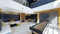 This contemporary entertainment lounge is designed with both socializing and recreation in mind. The space is immediately distinguished by its soaring double-height ceilings, which are complemented by a mezzanine level enclosed with sleek glass railings, adding depth and an open, airy atmosphere throughout.

The main floor is anchored by a full-length shuffleboard table, making it a focal point for recreational activity. Adjacent to it, a wall-mounted large-screen TV creates a dedicated viewing area, ideal for watching sports or entertainment. Warm wood-toned structural columns and slatted ceiling details introduce natural texture, balancing the otherwise contemporary, dark-accented interior palette.

A built-in dining nook with a solid wood table and bench seating offers an intimate gathering spot, framed by a decorative shelving unit displaying curated wall art and accessories. Pendant lighting with sleek white fixtures hangs above, providing focused illumination. In the lounge area, casual seating with soft furnishings and playful accent pieces in green and white tones provide a relaxed counterpoint to the more active zones. Light-toned flooring throughout ties the various areas together cohesively.