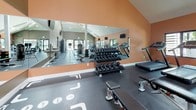 This fitness center is thoughtfully designed to accommodate a wide range of workout styles and fitness goals. The space is divided into distinct functional zones, each optimized for specific training activities. The cardio area features a row of treadmills, elliptical machines, and a rowing machine, all positioned along warm terracotta-painted walls with windows that invite natural light and outdoor views. Multiple wall-mounted flat-screen televisions are placed throughout for entertainment during workouts. The free weights section is equipped with a full rack of dumbbells and adjustable benches, complemented by a large floor-to-ceiling mirror that enhances the sense of space and allows for form checking. A separate functional training zone houses a dual cable machine system, colorful kettlebells, and additional benches on durable rubber flooring marked with training guides. The main area boasts soaring vaulted ceilings with exposed beams and skylights, creating an airy, open atmosphere. Recessed lighting ensures consistent illumination throughout. A dedicated spin room with cycling bikes offers a more focused cardio experience. Practical amenities include dual water fountains and a waste station. The overall design combines neutral taupe and terracotta tones, contributing to an energizing yet focused workout environment.