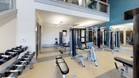 This well-appointed fitness center spans two floors, offering a thoughtfully designed environment for a full range of workout activities. The main floor immediately impresses with soaring double-height ceilings and an abundance of natural light streaming through large, multi-pane windows positioned at both lower and upper levels. Bold accent walls in a deep teal blue add a dynamic, energizing atmosphere, complemented by full-length mirrors that visually expand the space and allow for proper form checks during exercise.

The main workout floor is equipped with a wide array of strength-training machines, including cable systems, weight benches, and functional trainers, alongside a well-stocked dumbbell rack. Multiple flat-screen televisions are mounted at elevated positions throughout, providing entertainment during workouts. The flooring is a durable speckled rubber material, ideal for high-traffic fitness use, and a ceiling fan aids in air circulation. Dedicated restroom facilities are conveniently located on this level as well.

The second floor, accessible via an interior staircase with a sleek metal handrail, offers additional workout space featuring striped carpet tile flooring and cardio equipment, including a treadmill. This upper level provides a more intimate setting, with security and climate control elements integrated discreetly into the clean, white walls. Together, both levels deliver a comprehensive and functional fitness experience.