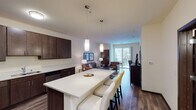 This single-floor residence is designed with a modern, open-concept layout that connects the kitchen, dining, and living areas into one cohesive space. The kitchen is outfitted with dark espresso cabinetry extending to the ceiling, stainless steel appliances including a refrigerator, range, microwave, and dishwasher, and a light quartz countertop. A large central island with pendant lighting above serves as both a prep space and casual dining area with bar seating.

The living area flows naturally from the kitchen and features hardwood-style flooring, recessed lighting, and a sliding glass door that opens to a private balcony with outdoor views. The space accommodates a full seating arrangement and a dedicated workspace or media area.

Two bedrooms are carpeted and each receives natural light through windows with blinds. Both are well-proportioned, offering room for full bedroom furniture sets.

The bathroom features dark wood vanity cabinetry, a quartz-style countertop, a large mirror, wood-look tile flooring, and a separate walk-in shower.

A dedicated in-unit laundry room houses a stacked washer and dryer unit alongside open shelving, providing convenient everyday functionality within the residence.