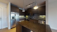 This contemporary single-floor residence showcases a thoughtfully designed open layout with warm hardwood-style flooring that flows seamlessly throughout the main living areas. The kitchen is a standout feature, boasting rich dark cabinetry that extends to the ceiling for ample storage, deep granite countertops, and a full suite of stainless steel appliances including a French door refrigerator with ice maker, range, microwave, and dishwasher. A tiled backsplash in a subtle green-grey tone adds texture and depth, while a central island provides additional prep and dining space. Pendant lights and a track lighting system illuminate the kitchen with a warm, layered glow. The open-concept design connects the kitchen directly to the living area, creating an expansive, airy feel ideal for everyday living and entertaining. The bedroom is bright and inviting, featuring large windows with horizontal blinds that flood the room with natural light and offer views of greenery outside, complemented by a ceiling fan for year-round comfort. The bathroom is finished with wood-toned tile in the tub surround, light stone-look floor tiles, a dark vanity with granite countertops, and clean, modern fixtures throughout. Overall, the space combines contemporary design with functional details for a polished, comfortable living environment.