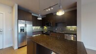 This contemporary single-floor residence showcases a thoughtfully designed open layout with warm hardwood-style flooring that flows seamlessly throughout the main living areas. The kitchen is a standout feature, boasting rich dark cabinetry that extends to the ceiling for ample storage, deep granite countertops, and a full suite of stainless steel appliances including a French door refrigerator with ice maker, range, microwave, and dishwasher. A tiled backsplash in a subtle green-grey tone adds texture and depth, while a central island provides additional prep and dining space. Pendant lights and a track lighting system illuminate the kitchen with a warm, layered glow. The open-concept design connects the kitchen directly to the living area, creating an expansive, airy feel ideal for everyday living and entertaining. The bedroom is bright and inviting, featuring large windows with horizontal blinds that flood the room with natural light and offer views of greenery outside, complemented by a ceiling fan for year-round comfort. The bathroom is finished with wood-toned tile in the tub surround, light stone-look floor tiles, a dark vanity with granite countertops, and clean, modern fixtures throughout. Overall, the space combines contemporary design with functional details for a polished, comfortable living environment.