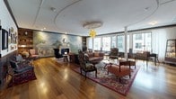 This elegantly designed single-floor space offers a compelling blend of classic craftsmanship and modern sensibility. The main lounge area is anchored by a sweeping, hand-painted landscape mural that spans an entire wall, evoking a sense of history and grandeur. A decorative white marble fireplace sits beneath the mural, adding warmth and architectural distinction. Rich hardwood flooring runs throughout, complementing the dark navy wainscoting that lines the walls and contributes to the space's refined, club-like atmosphere. The lounge is furnished with an eclectic mix of vintage and contemporary seating — including tufted leather sofas, ornate upholstered chairs, and sculptural accent pieces — arranged over vibrant patterned area rugs. A striking gold pendant light hangs from a recessed ceiling medallion, serving as a central focal point. Built-in bookshelves and a decorative globe add an intellectual, worldly character. A gallery-style hallway features a dense arrangement of framed artwork and prints. Floor-to-ceiling windows along one side of the space flood the interior with natural light and offer views of the surrounding urban landscape. An adjoining area with banquette seating and marble-top tables provides an intimate setting, completing this richly layered and thoughtfully curated environment.