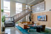 Located at 868 S Fifth St in San Jose, California, this impressive residential lobby offers a welcoming and thoughtfully designed communal space. The interior is defined by soaring double-height ceilings that flood the area with natural light through expansive floor-to-ceiling windows, creating an open and airy atmosphere throughout.

The polished terrazzo flooring at the entry transitions seamlessly into richly hued teal carpet that anchors the central lounge zones. A striking feature of the space is its curated collection of artwork, including bold geometric murals, a woven fiber wall hanging, and abstract canvas pieces that lend the lobby a gallery-like character.

Seating is arranged across several distinct areas to encourage both social interaction and quiet relaxation. Tan leather sofas, modern accent chairs, and small round tables are thoughtfully positioned alongside communal dining-height tables and bar seating for more active use. A wall-mounted display screen serves as an informational hub for residents.

Natural wood wall paneling adds warmth to the otherwise contemporary palette of whites and neutrals, while a sculptural multi-arm pendant light fixture provides a dramatic focal point overhead. A sleek mailroom partition and a compact service counter with cabinetry and a sink near the entrance add practical functionality. Large planters with lush greenery introduce organic texture, completing this dynamic and inviting community space.