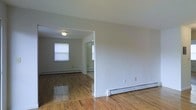 This single-floor space offers a clean, functional layout with a cohesive design carried throughout. The interior is defined by warm, polished hardwood floors that extend seamlessly from room to room, reflecting light and adding a sense of natural warmth to the space. Neutral-toned walls in a soft gray-white palette create a bright, versatile backdrop that adapts well to a variety of furnishing styles. The open layout connects multiple areas through wide passageways, allowing for easy flow between the main living space and adjoining rooms. Natural light enters through a window in the adjacent room, further brightening the interior. Baseboard heating units are present along the lower walls, providing efficient climate control throughout the space. The ceilings are smooth and finished with flush-mount lighting, contributing to a tidy, understated aesthetic. White trim and baseboards frame the rooms cleanly, adding a crisp architectural detail that complements the overall neutral tone. The space presents a straightforward, well-maintained interior well-suited for comfortable everyday living.