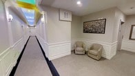 This single-floor space presents a well-appointed interior characterized by classic architectural details and a polished, neutral aesthetic. The hallway is a standout feature, lined with crisp white wainscoting panels that extend along the walls, adding a refined, traditional touch to the overall design. The walls are finished in soft, warm neutral tones that contribute to a calm and welcoming ambiance throughout.

A cozy seating alcove is integrated into the corridor, furnished with two upholstered barrel-back chairs accented with nailhead trim, offering a comfortable resting area. A framed artwork piece is displayed prominently on the wall above the seating area, lending a touch of sophistication to the space.

The flooring transitions between patterned carpet in the main corridor and a lighter, neutral carpet in the seating nook, providing visual distinction between functional and relaxation areas. Recessed ceiling lighting provides clean, even illumination, while wall-mounted sconces add a warmer, accent glow along the hallway. A glimpse of colorful decorative elements is visible at the far end of the corridor, suggesting additional vibrant design features deeper within the space. Overall, the interior conveys a professional yet inviting character, well-suited for hospitality or institutional use.