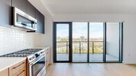 This contemporary single-floor residence combines sleek design with functional living. The kitchen is a standout feature, outfitted with stainless steel appliances including a gas range, built-in microwave, French door refrigerator, and dishwasher. Cabinetry contrasts warm wood-toned lower cabinets with dark upper cabinets in teal and charcoal finishes, complemented by a white tile backsplash and light stone countertops. Light hardwood flooring runs throughout the entire unit, lending warmth and continuity to each space. Floor-to-ceiling sliding glass doors in both the main living area and a bedroom open directly onto a private balcony, flooding the interior with natural light and offering sweeping elevated views of the surrounding cityscape and greenery. The bedrooms are bright and minimal, featuring clean white walls and sliding closet doors with built-in rod storage. The overall design aesthetic is clean and modern, with a neutral palette of whites, natural wood, and matte black accents throughout. The open layout and generous glazing create an airy, light-filled atmosphere, while the elevated position of the unit enhances the sense of openness and connection to the urban environment beyond.