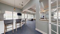 Located in Integra Woods in Palm Coast, Florida, this single-floor community clubhouse offers a thoughtfully designed gathering space with a cohesive coastal aesthetic throughout. The interior is finished in soft sage-green tones complemented by crisp white trim, coffered and tray ceilings with crown molding, and deep blue carpeting that anchors the open layout.

The entrance leads into a spacious open area that seamlessly connects the kitchen, lounge, and game zones. The kitchen features white shaker-style cabinetry, granite countertops, a center island with bar seating and pendant lighting, tile flooring, and stainless steel appliances — ideal for community events and gatherings. A wall-mounted television adds functionality to the kitchen and bar area.

The living and game area is generously sized, accommodating a pool table, shuffleboard table, and a dedicated gaming nook with an arcade-style table. Comfortable lounge seating is arranged throughout for relaxed socializing. Multiple wall-mounted televisions serve the entertainment zones.

Natural light pours in through an abundance of white-trimmed windows dressed with classic plantation-style blinds, offering views of the surrounding greenery. Stylish pendant and chandelier lighting enhances the ambiance, while recessed lighting ensures the entire space is well-illuminated. The overall design balances comfort and functionality for an engaging community experience.