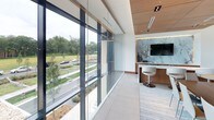 This single-floor space exemplifies contemporary design through a thoughtful combination of high-end materials, clean lines, and a seamless connection between interior and exterior environments. Floor-to-ceiling glass curtain walls form a defining architectural feature, flooding the interior with natural light while offering expansive views of the surrounding greenery and landscaped grounds.

The main gathering area is anchored by a substantial dark walnut conference or dining table surrounded by sleek upholstered chairs, positioned beneath a sculptural cluster pendant light fixture that serves as a striking focal point. Warm wood panel ceilings contrast beautifully with crisp white walls and large-format light tile flooring, creating a balanced interplay of textures throughout.

A built-in display niche with warm wood framing showcases curated sculptural artwork, adding an artistic dimension to the space. Adjacent areas feature a work counter with a stainless steel sink, walnut cabinetry, and light stone countertops, providing functional utility without compromising aesthetic cohesion.

A secondary lounge or work area includes a wall-mounted display screen set against a dramatic stone-textured accent wall, accompanied by a modern swivel stool and compact built-in workspace, reinforcing the space's versatile, professional character throughout.