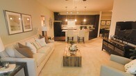 This single-floor residence showcases an open-concept layout that connects the living area and kitchen into one cohesive, functional space. The living area features plush, neutral-toned carpeting and is anchored by a comfortable cream sofa accented with decorative pillows. A rustic reclaimed wood coffee table serves as a standout centerpiece, adding textural contrast to the soft surroundings. Warm ambient lighting from a ceiling fan and a floor lamp enhances the cozy atmosphere.

The kitchen is defined by rich dark cabinetry that extends to the upper walls, creating a bold contrast against the light countertops and a classic white subway tile backsplash. A central island with seating for multiple occupants serves as both a prep area and casual dining spot, illuminated by a trio of pendant lights that add a refined, modern touch. Black appliances complement the dark cabinetry for a cohesive aesthetic. Track lighting overhead ensures the kitchen remains well-lit and practical. A glimpse of a separate dining area is visible to the right, suggesting additional dedicated space for entertaining. Overall, the space balances warmth, functionality, and modern design elements cohesively.