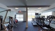 This well-appointed fitness center offers a clean, contemporary design with a functional layout suited for a comprehensive workout experience. The space is anchored by a row of treadmills lined along one wall, each positioned to face a wall-mounted television, making cardio sessions both engaging and comfortable. Large, floor-to-ceiling windows flood the room with natural light while offering open views of the surrounding outdoor scenery, creating an energizing atmosphere for exercise.

On the opposite side of the space, a dedicated free weights area features a rack of medicine balls, dumbbells, and a weight bench, providing ample options for strength training. Structural columns are integrated seamlessly into the layout without disrupting the flow of the space. The flooring consists of durable, patterned carpet designed to withstand high-traffic use while providing comfort underfoot.

Recessed circular ceiling lights supplement the abundant natural light, ensuring the space remains well-lit at all hours. The cool, neutral color palette — featuring grey tones on the walls, ceiling, and flooring — contributes to a focused and professional ambiance. Ventilation is provided through ceiling-integrated HVAC systems, keeping the environment comfortable during intense workouts. Overall, the space strikes a balance between practicality and modern design.