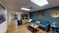 Located at 5800 Maudina Ave in Nashville, TN, this single-floor commercial space is thoughtfully designed to blend functionality with a warm, creative atmosphere. Throughout the space, light hardwood flooring, drop ceilings with recessed panel lighting, and bold teal accent walls create a cohesive and inviting environment.

The lounge area serves as a central gathering point, furnished with a tufted teal sofa, white accent chairs, a navy blue leather chair, and a wood-and-metal coffee table. A decorative gold sunburst mirror and vibrant guitar-themed artwork adorn the walls, adding a distinctly Nashville flair. A rustic wood shelving unit provides functional display storage.

Adjacent to the lounge, a dedicated group seating area features two large oval tables with ornate resin tops, surrounded by chairs and framed by colorful musical instrument artwork on teal walls — ideal for group meetings or collaborative sessions.

Private workstation areas offer focused workspace, each equipped with a desk, computer setup, and contemporary artwork. One compact office features a modern pod-style chair and a glass accent table, balancing practicality with design.

The kitchen is well-appointed with white shaker cabinetry, dark countertops, a stainless steel refrigerator, microwave, sink, and coffee station, providing full break room functionality. A secondary door in the kitchen offers convenient exterior access.