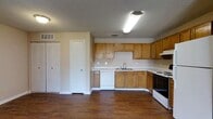 This single-floor space offers a practical and well-organized layout designed for comfortable everyday living. The kitchen serves as a central feature, showcasing warm honey-toned wood cabinetry that extends along the walls, providing generous storage and counter space. A double-basin sink sits beneath overhead cabinets, complemented by a white dishwasher and an electric range with a built-in exhaust hood. The light-colored countertops offer a clean contrast against the rich, dark hardwood-style flooring that extends throughout the space, adding warmth and a sense of continuity. A textured ceiling with recessed ventilation and fluorescent lighting ensures the kitchen area is well-lit and functional. Adjacent to the kitchen, a closet with bifold doors adds to the storage options within the space. A secondary door nearby provides additional access or utility. Neutral wall tones throughout keep the atmosphere bright and versatile, making the space easy to personalize. Overall, the layout is efficient and straightforward, offering a welcoming environment with durable finishes suited for practical, day-to-day use.