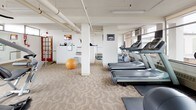 Located at 34 Carleton St in Cambridge, MA, this fitness center offers a well-equipped exercise space designed to accommodate a variety of workout routines. The room features patterned carpeting throughout and exposed overhead piping, lending an industrial character to the otherwise clean, white-painted interior. Structural columns punctuate the open floor plan, which is organized to maximize usable workout space.

The cardio zone is lined with multiple treadmills and elliptical machines positioned along large windows that frame views of the surrounding Cambridge and waterfront cityscape, allowing natural light to fill the space during workouts. A multi-station strength training machine and additional resistance equipment are positioned toward the center and perimeter of the room, offering users options for full-body conditioning. Stability balls and a ballet barre add functional training versatility.

A large wall-mounted mirror visually expands the space and reflects the natural light from the windows. A small utility counter area near the entrance provides a convenient spot for essentials. The fitness center is served by perimeter heating units, ensuring comfort year-round. Overall, the space delivers a functional and practical environment for daily exercise.
