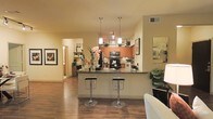 This single-floor residence showcases an open-concept layout that seamlessly connects the kitchen and living areas. The kitchen is a central highlight, featuring warm-toned wood cabinetry, dark granite countertops, and stainless steel appliances including a microwave and range. A peninsula-style breakfast bar extends into the living space, accommodating two modern chrome bar stools, making it ideal for casual dining or entertaining. Pendant lighting fixtures hang above the bar, providing focused task lighting while adding a contemporary design touch. The flooring throughout is rich hardwood, lending warmth and continuity across the open floor plan. Framed artwork adorns the walls, contributing to the curated, welcoming ambiance of the space. A hallway visible in the background suggests additional rooms beyond the main living area. The overall aesthetic is modern yet inviting, with a neutral warm color palette complemented by natural greenery and decorative floral arrangements that add life and character to the interior.