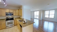 This single-floor, 1-bedroom, 1-bathroom residence offers a thoughtfully designed open-concept layout that seamlessly connects the kitchen and living areas. The kitchen is outfitted with light maple cabinetry, granite countertops, stainless steel appliances — including a range and built-in microwave — and a peninsula with an integrated sink that doubles as a breakfast bar, providing both functionality and a natural division between cooking and living spaces. The flooring transitions from tile in the kitchen to warm hardwood-style flooring throughout the main living area, adding visual continuity and a sense of warmth. The spacious living area is bathed in natural light, thanks to a large floor-to-ceiling sliding glass door that opens onto a private balcony, offering views of the surrounding area. The bright, neutral wall tones throughout enhance the airy and open ambiance of the home. The overall design balances modern finishes with practical living, making this a comfortable and stylish space suited for everyday living.