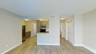 This single-floor residence offers a clean, modern layout designed for comfortable everyday living. The open-concept design seamlessly connects the kitchen and main living area, creating a bright and functional space. The kitchen is a standout feature, equipped with stainless steel appliances including a refrigerator, microwave, and range, all complemented by gray cabinetry, a tile backsplash, and a light granite-topped peninsula that serves as both a prep area and casual dining space. Track lighting overhead ensures the kitchen is well-lit and practical. Light hardwood-style flooring runs throughout the main living areas, lending a cohesive and contemporary feel to the interior. The neutral white and gray color palette keeps the space feeling open and airy. A hallway visible from the main area leads to additional rooms, including what appears to be bedrooms and a bathroom, each accessed through classic paneled doors. The layout is efficient and thoughtfully organized, with ample closet access visible along the corridor. Overall, the space presents a tidy, move-in-ready environment with a modern aesthetic that balances style and functionality.