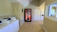 This single-floor communal laundry facility offers a practical and functional space designed to serve multiple users efficiently. The room is characterized by its notable vaulted ceiling, which lends a sense of openness and airiness that is uncommon in utility spaces of this kind. The walls are finished in a neutral, light green tone that contributes to a clean and welcoming atmosphere.

The laundry area is equipped with both stacked and top-load washer and dryer units, providing flexibility for users with varying laundry needs. The machines are arranged along the walls to maximize the open floor space at the center of the room. Light-toned ceramic tile flooring runs throughout, offering durability and ease of maintenance.

A prominently placed Coca-Cola vending machine adds a convenient amenity for users during their visit. A thermostat on the wall suggests climate control is available, ensuring comfort year-round. A doorway visible toward the back of the space leads to an additional room or utility area, hinting at further functional square footage beyond the main laundry room. Signage on the wall clearly identifies the space as a laundry facility. Overall, the space balances utility with a tidy, well-maintained environment.