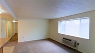 This single-floor space presents a clean and functional layout designed for comfortable everyday living. The main living area features wall-to-wall carpeting in a neutral beige tone, providing a soft and understated foundation throughout the room. The walls are finished in a light cream color, contributing to a bright and open atmosphere. A textured ceiling adds a subtle visual element overhead, characteristic of classic residential construction. Natural light filters into the space through a wide horizontal window fitted with blinds, allowing for adjustable brightness and privacy. A wall-mounted HVAC unit is positioned beneath the window, offering convenient in-unit climate control for year-round comfort. Electrical outlets are evenly distributed along the walls, ensuring practical utility throughout the room. The overall ambiance of the space is modest and neutral, providing a blank canvas that is well-suited for personalization with furnishings and décor. The straightforward design prioritizes functionality, making it an accessible and easy-to-maintain living environment.