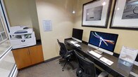 This single-room business center is designed to provide a functional and efficient workspace for guests. The room features two workstations arranged along a continuous built-in desk that runs along one wall, each equipped with an Apple iMac computer, keyboard, and mouse. Ergonomic task chairs accompany each station, ensuring comfort during use. The space is finished with neutral, warm-toned walls and carpeted flooring, contributing to a quiet, focused atmosphere. Pendant lighting hangs from the ceiling, providing targeted illumination over the work area. A framed black-and-white photograph is mounted on the wall above the desk, adding a subtle decorative element. To the left, a small storage cabinet and what appears to be a printer or additional equipment offer supplementary office functionality. A posted "Business Center Guidelines" sign on the wall ensures guests are informed of usage policies. Overall, the space is compact yet well-organized, prioritizing utility and ease of use for travelers or guests requiring access to computing resources.