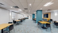Located at 1001-1003 Easton Road in Willow Grove, Pennsylvania, this single-floor commercial space offers a generously sized, open-plan layout ideal for gatherings, meetings, or community use. The interior is defined by a distinctive patterned carpet in shades of blue and tan, which anchors the main seating area and complements the clean, neutral white walls throughout.

The main hall is furnished with multiple square tables and modern chairs arranged in a flexible, clustered configuration, accommodating a variety of group sizes. Bold teal accent columns and a striking yellow wood-paneled media wall—complete with a wall-mounted flat-screen television—provide visual interest and define functional zones within the open space. Colorful pop-art style prints adorn the walls, adding energy and character to the environment.

A built-in counter bar with teal upholstered stools serves as a dedicated lounge or refreshment area, finished with a clean white countertop and white tile flooring that contrasts the carpeted main zone. Plantation shutters along multiple windows allow for controlled natural light throughout the space. A glass-partitioned entry area with a private meeting room or office is accessible via double glass doors, and restroom facilities are conveniently located off the main corridor. Recessed and panel lighting ensures consistent, well-lit conditions throughout.