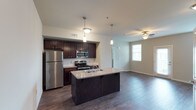 This single-floor space showcases an open-concept layout that integrates the kitchen and living area into one cohesive environment. The kitchen is a standout feature, equipped with stainless steel appliances including a refrigerator, range, and built-in microwave. Rich, dark espresso cabinetry lines the walls, offering generous storage, while light granite countertops provide a striking contrast. A central kitchen island with a built-in sink and matching granite surface serves as both a functional workspace and a natural gathering point. Pendant lighting fixtures hang above the island, adding focused illumination and a touch of modern style. Dark hardwood flooring extends throughout, lending warmth and continuity between the kitchen and the adjoining living area. The living space is bright and open, benefiting from a window that allows natural light to filter in. A ceiling fan adds to the comfort of the room. The overall aesthetic is clean and contemporary, with a neutral color palette that complements the dark cabinetry and flooring, creating a balanced and inviting atmosphere.