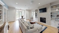 This single-floor clubhouse offers a well-appointed community gathering space designed with a clean, coastal-inspired aesthetic. The main lounge area is bright and airy, benefiting from abundant natural light streaming through multiple French doors and large windows that offer views of the surrounding outdoor areas. Light hardwood flooring runs throughout, complementing the neutral white walls and soft furnishings. A striking stone-surround fireplace serves as the focal point of the lounge, flanked by built-in shelving and cabinetry, and topped with a wall-mounted television — ideal for both relaxed socializing and community events. Comfortable seating, including a generous sofa and accent chairs, is arranged around a central coffee table atop a navy blue area rug, creating an inviting conversational layout. The kitchenette is thoughtfully designed with white shaker-style cabinetry, quartz countertops, a stainless steel refrigerator, an undermount sink, and a dedicated beverage station complete with a coffee machine and cupped drink supplies. Soft blue accent walls in the kitchen area add a touch of warmth and coastal character. Additional built-in shelving and a round mirror contribute to the space's polished, functional design. French doors provide seamless access to an outdoor patio, extending the usable space for gatherings.