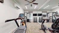 Located in Dahlonega, Georgia, this fitness center offers a compact yet thoughtfully outfitted exercise room designed to support a well-rounded workout routine. The space features crisp white walls accented by crown molding, neutral-toned carpet flooring, and bright linear lighting complemented by a ceiling fan for added air circulation. A wall-mounted mini-split system ensures year-round climate control and comfort during workouts.

The room is equipped with a treadmill and a stationary bike for cardiovascular training, alongside a multi-function cable machine ideal for strength and resistance exercises. A complete dumbbell rack with a full range of weights and a colorful medicine ball wall rack provide versatile options for functional fitness. A flat-screen television is mounted on the wall, offering entertainment during exercise sessions.

Full-length mirrors line one wall, visually expanding the space while allowing users to monitor their form. Natural light filters through windows dressed with fabric roman shades, contributing to an inviting atmosphere. A second entry door and posted fitness center rules and guidelines reflect the facility's organized, community-oriented design. The overall ambiance is clean, modern, and purposeful — a well-appointed fitness resource for residents in the Dahlonega area.