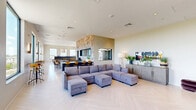 Located at 39 Trotter Rd in Weymouth, MA, this contemporary amenity space delivers an impressive combination of interior comfort and outdoor living across a generously sized single floor. The interior lounge area is anchored by a striking feature wall clad in reclaimed wood, housing a built-in fireplace and mounted TV, flanked by leather-upholstered benches and open display shelving. Light herringbone-patterned hardwood flooring runs throughout, complementing the bright, neutral palette and recessed lighting overhead. Floor-to-ceiling windows flood the space with natural light and frame sweeping views of the surrounding Weymouth skyline and treetops. A textured stone accent wall defines the open-concept bar and dining area, complete with a dark countertop island and amber bar seating. Adjacent to the lounge, a dedicated game room features a ping pong table, booth-style seating with navy and orange upholstery, and casual high-top tables. The rooftop terrace extends the living space outdoors with large-format tile pavers, a built-in wood bench seating area centered around a sleek gas fire table, decorative landscaping, a mounted outdoor TV, and festoon string lighting that creates an inviting atmosphere for evening gatherings, all beneath open skies with panoramic views.