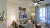 This single-floor space showcases a well-appointed living area designed with both comfort and style in mind. The living room serves as the centerpiece, featuring a light gray sofa accented with decorative throw pillows, anchored by a patterned area rug that adds warmth and visual interest to the floor. A large-scale abstract canvas painting in blues, whites, and earth tones dominates the main wall, serving as a bold focal point that elevates the room's aesthetic.

A ceiling fan with an integrated light fixture is centrally mounted, providing both air circulation and ambient lighting. The space is further illuminated by a pair of floor and table lamps, creating a layered lighting scheme suited for various settings. Sheer, textured curtains frame a window along one wall, filtering natural light into the room while maintaining a sense of privacy.

Crown molding along the upper walls adds a refined architectural detail, contributing to the overall polished character of the space. The neutral wall tones provide a versatile backdrop, complementing the mix of contemporary furnishings and decorative elements throughout the room.