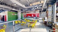 This lively fast-casual restaurant is designed with a bold, industrial-chic aesthetic that immediately captures attention. The single-floor layout encompasses a generously sized dining area and a fully equipped back-of-house kitchen. Guests enter through a street-facing glass entrance, greeted by a striking living moss wall emblazoned with the "I ♥ mac &" logo, setting a playful and welcoming tone.

The dining area features polished concrete floors, an exposed ceiling with visible ductwork, and hanging Edison bulb pendants suspended from reclaimed wood beams. Vibrant yellow metal chairs and dark wood-top tables are arranged throughout, complemented by a window-side counter seating area with bar-height stools offering street views. Every wall is adorned with energetic, hand-drawn-style illustrated murals on dark backgrounds, with one prominent wall clad in bold red subway tile behind the service counter.

The service counter itself is finished with corrugated metal paneling and a dark countertop, providing a functional and visually striking ordering experience. Natural light pours in through large floor-to-ceiling windows, enhancing the open and energetic ambiance. The back-of-house kitchen is a fully operational commercial space, outfitted with stainless steel sinks, industrial shelving, and dedicated storage, reflecting the restaurant's operational efficiency.