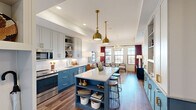 Located at 98 South River Drive in Tempe, Arizona, this thoughtfully designed one-bedroom, one-bathroom residence offers a refined living experience within a efficiently utilized floor plan. The open-concept layout seamlessly connects the kitchen and living room, creating a spacious and inviting atmosphere throughout.

The kitchen is a standout feature, boasting two-toned cabinetry with light gray uppers and striking blue lowers, complemented by a large quartz-topped island with built-in shelving and seating. Stainless steel appliances, white subway tile backsplash, and bold brass pendant lighting complete this well-appointed culinary space.

The living room is bathed in natural light from large gridded windows dressed with dramatic burgundy drapes. A sculptural chandelier anchors the room, which features warm hardwood-style flooring that flows throughout the home.

The bedroom is a serene retreat with a ceiling fan, natural light from a generous window, and ample room for a full furniture arrangement. The bathroom offers a long vanity with granite countertops, white cabinetry with gold hardware, large-format tile flooring, and a full tub-shower combination. Every room reflects a cohesive, upscale aesthetic with carefully considered design details.