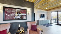 Located at 115 E 5th Ave in Columbus, Ohio, this rooftop amenity space combines vibrant interior design with an expansive outdoor deck to create a dynamic gathering environment. The interior lounge is immediately eye-catching, featuring a boldly striped yellow and gray ceiling with recessed lighting, herringbone-patterned wood-look flooring, and an exposed dark brick accent wall adorned with large-scale artwork. A built-in wet bar with sage-green cabinetry, a stainless steel sink, and a mounted flat-screen TV provide functional elegance along one wall. A cozy seating nook with plush velvet armchairs and a patterned area rug adds a warm, social atmosphere. A standout architectural feature is the oversized industrial glass garage door that opens to the outdoor deck, seamlessly blending the interior and exterior spaces. Along the interior perimeter, a built-in wood bench with cushions and a window-facing bar counter with wicker stools invite guests to take in the surrounding cityscape. The outdoor rooftop deck extends the entertaining area with long communal wood dining tables, bar-height seating, and multiple built-in grilling stations, all framed by a modern gray perimeter wall. Together, these spaces offer a well-appointed, design-forward venue suited for social gatherings and events.