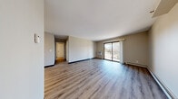 This 1-bedroom, 1-bathroom unit offers a functional and comfortable layout across a single floor. The open living area is the centerpiece of the space, featuring light-toned luxury vinyl plank flooring, neutral-toned walls, and a wall-mounted air conditioning unit. A large sliding glass door leads to a private balcony, flooding the room with natural light and offering pleasant outdoor views of surrounding trees and greenery. A hallway connects the main living area to the bedroom and bathroom, providing a clear and organized flow throughout the unit. The bedroom is a comfortable, well-proportioned room finished with soft carpeting and a window fitted with blinds that allows for ample natural light while maintaining privacy. Baseboard heating runs along the walls, ensuring efficient warmth throughout. The bathroom features light vinyl plank flooring, a white vanity with dark granite countertop, a tub-shower combination, and built-in shelving, offering both style and practicality. The kitchen, visible from the 3D overview, is efficiently designed to maximize the available space. Overall, the unit presents a clean, move-in-ready aesthetic with consistent, modern finishes throughout.
