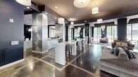 Located at 3109 Douglas Avenue in Dallas, TX, this upscale amenity space delivers a refined, contemporary experience across a single, expansive floor. The layout encompasses three distinct living areas, a connecting hallway, and a reception zone, all unified by a cohesive modern aesthetic. Polished concrete floors with geometric inlay patterns anchor the main gathering areas, while plush marbled-pattern carpeting defines the softer lounge zones. Dramatic crystal chandelier pendants cascade from elevated ceilings throughout, serving as striking focal points in multiple rooms. Floor-to-ceiling window walls flood the space with natural light and frame views of the mature trees lining Douglas Avenue, creating a seamless connection between interior and exterior. A game lounge features a billiards table and shuffleboard, set against bold dark walls and large glazed panels. A separate screening or group seating room accommodates numerous curved velvet chairs arranged around a vibrant large-scale abstract artwork, with track and recessed lighting setting an intimate mood. The reception area presents a sleek white high-top counter, sculptural wall art, and elegant drum pendant lights. Throughout, a carefully curated palette of charcoal, taupe, navy, and white, accented by metallic finishes and bold art pieces, establishes a polished, hospitality-inspired ambiance.